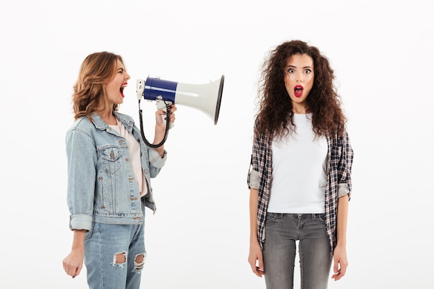 Photo gratuite choqué femme bouclée couvrant ses oreilles tandis que la deuxième fille lui criant avec mégaphone sur mur blanc