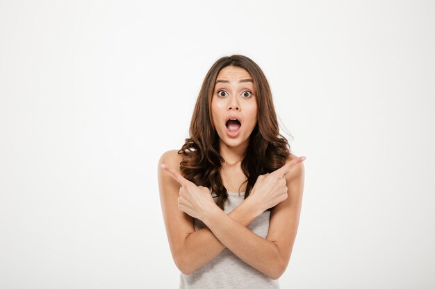 Choqué femme brune pointant vers les côtés et regardant la caméra sur gris