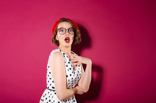 Choqué femme gingembre en robe et lunettes regardant la caméra sur rose