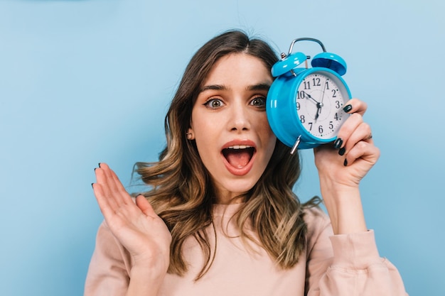 Choqué, femme, poser, à, bouche ouverte, matin, projectile studio, de, beau, bouclé, girl, à, horloge