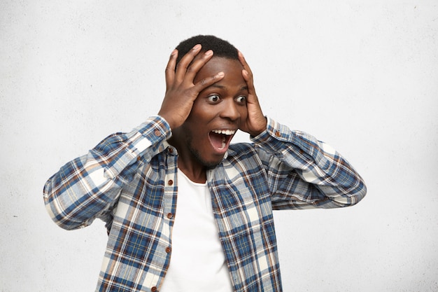 Photo gratuite choqué et hébété, jeune homme afro-américain en chemise à carreaux tenant la tête avec les deux mains regardant devant lui avec les yeux sortis et la bouche grande ouverte, ne peut pas croire sa chance, sa victoire ou son succès