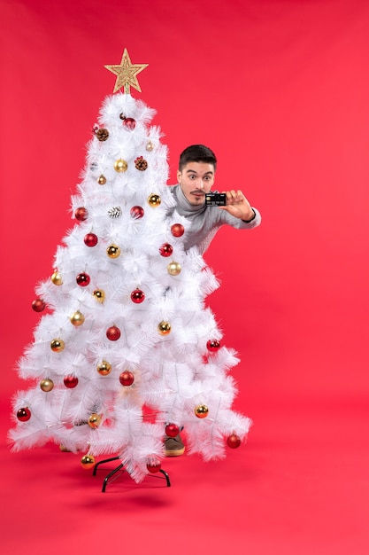 Choqué Jeune Adulte Dans Un Chemisier Gris Debout Derrière L'arbre De Noël Décoré Et Regardant Son Téléphone Sur Rouge