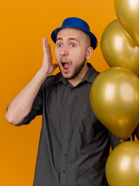 Choqué jeune beau mec de fête slave portant chapeau de fête tenant des ballons en gardant la main près de la tête à la droite isolé sur fond orange