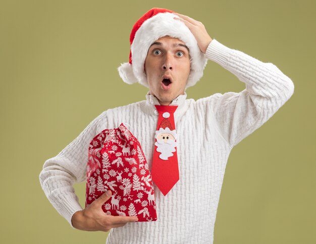 Choqué jeune beau mec portant chapeau de Noël et cravate de père Noël tenant le sac de Noël regardant la caméra en mettant la main sur la tête isolé sur fond vert olive