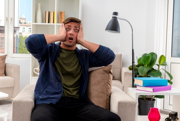 Choqué jeune bel homme blond est assis sur un fauteuil tenant la tête et regardant la caméra à l'intérieur du salon