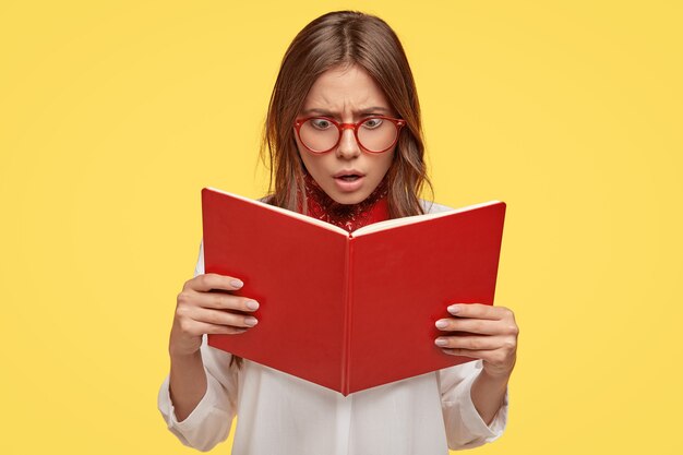 Choqué jeune brune avec des lunettes posant contre le mur jaune