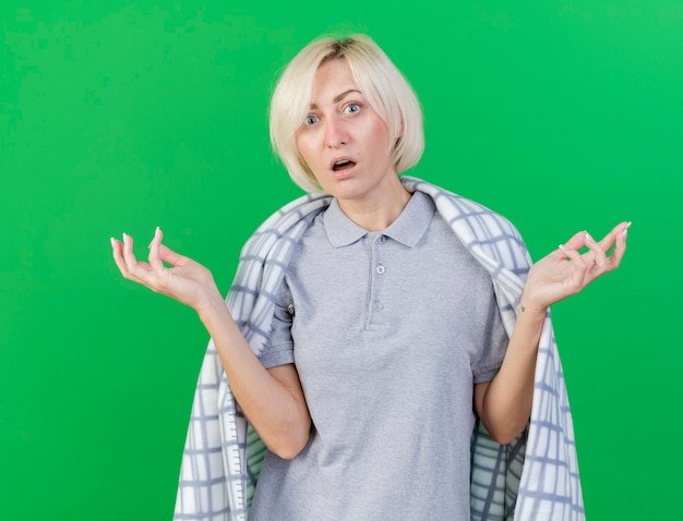 Photo gratuite choqué jeune femme slave malade blonde enveloppée dans un plaid tient les mains ouvertes isolé sur un mur vert avec espace copie