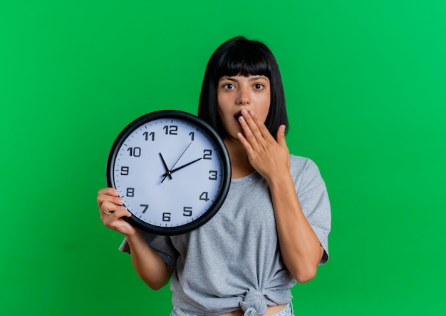 Choqué jeune fille caucasienne brune met la main sur la bouche et tient l'horloge