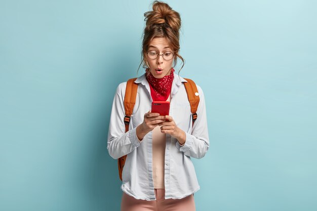 Choqué, jeune fille hipster regarde étonnamment le téléphone portable, reçoit un message inattendu, porte un sac à dos