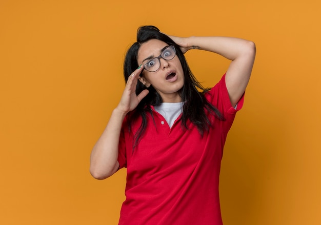 Choqué jeune fille de race blanche brune à lunettes optiques portant une chemise rouge tient la tête à la recherche d'isolement sur le mur orange