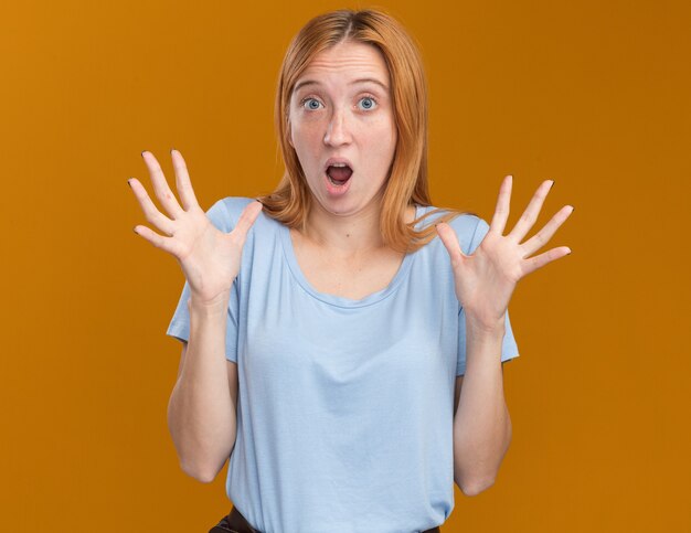 Choqué jeune fille rousse au gingembre avec des taches de rousseur debout avec les mains levées sur orange