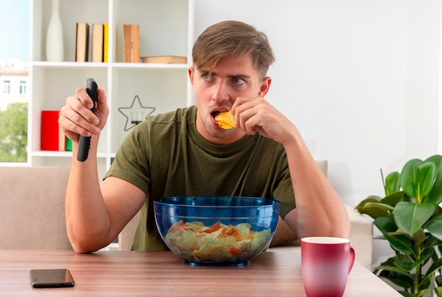 Choqué jeune homme beau blond est assis à table avec téléphone bol de chips et tasse tenant la télécommande du téléviseur et manger des chips à l'intérieur du salon