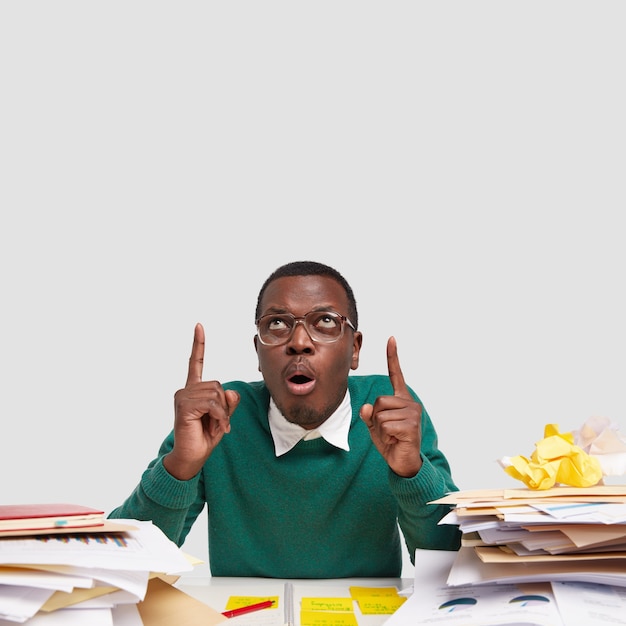 Choqué, jeune homme à la peau sombre pointe vers le haut avec les deux index, porte de grandes lunettes, entouré d'une pile de papiers