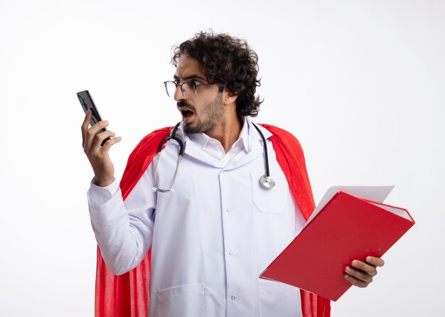Choqué jeune homme de super-héros caucasien à lunettes optiques portant l'uniforme de médecin avec manteau rouge et avec stéthoscope autour du cou détient le dossier et regarde le téléphone isolé sur le mur blanc