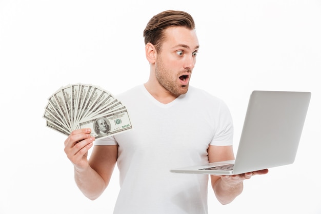 Choqué jeune homme tenant de l'argent à l'aide d'un ordinateur portable.