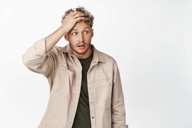 Choqué jeune homme tenant la main sur le front facepalm et regarde de côté le gros problème debout en détresse sur fond blanc