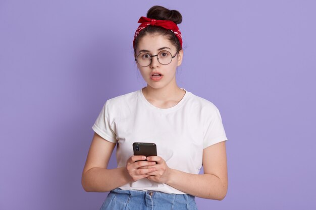 Choqué jeune jolie femme brune avec bandeau à la caméra étourdie, tenant le téléphone isolé sur l'espace lilas