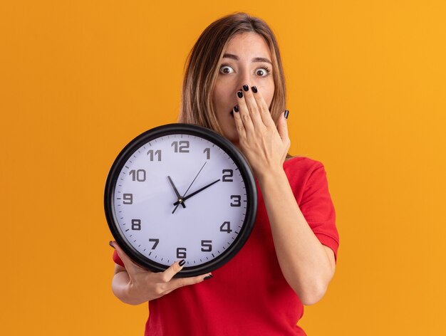 Choqué jeune jolie fille caucasienne met la main sur la bouche et tient l'horloge sur l'orange