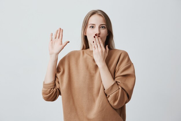 Choqué jeune mannequin avec des cheveux blonds teints directement étant perplexe, posant contre un mur gris avec des yeux embués, cachant sa bouche derrière sa main