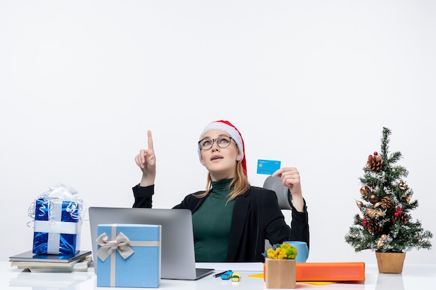 Choqué jolie femme avec chapeau de père Noël et portant des lunettes assis à une table cadeau de Noël et tenant une carte bancaire pointant au-dessus dans le bureau