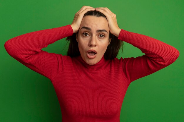 Choqué jolie femme tient la tête à deux mains isolé sur mur vert