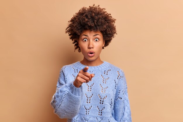 Choqué, sans voix, jeune femme regarde à l'avant et pointe l'index à l'avant porte un pull bleu d'hiver décontracté voit quelque chose d'horrible dans la distance pose contre le mur beige