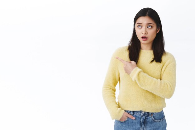 Choqué et sans voix, la jeune fille asiatique laisse tomber la mâchoire de l'étonnement bouche ouverte et regarde à gauche pointant du doigt une publicité choquante surprise debout fond blanc