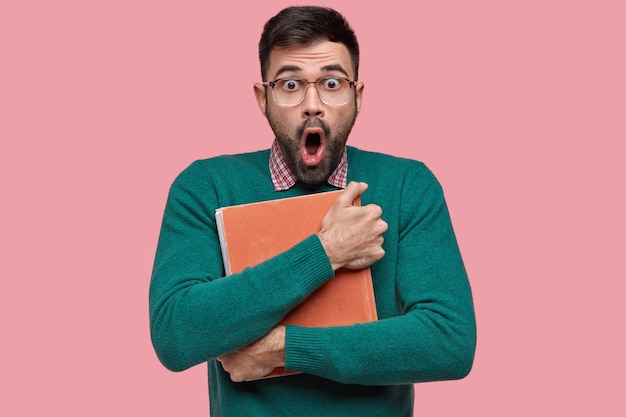 Photo gratuite choqué, surpris, jeune homme mal rasé, porte un livre vintage près de la poitrine, regarde avec des yeux buggés, a peur, porte des lunettes