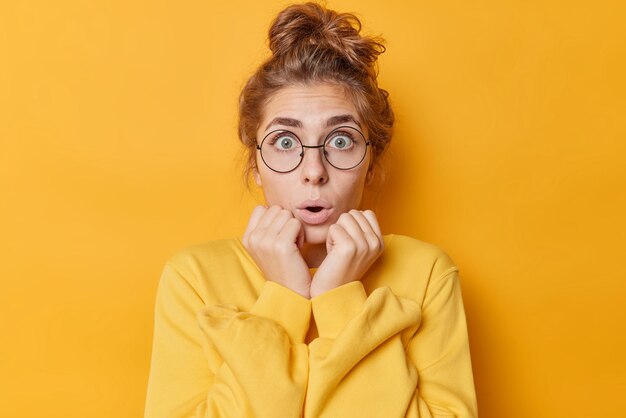 Choquée et étonnée, une jeune femme européenne halète et garde les mains sur les joues, les regards surpris garde la bouche ouverte porte des lunettes rondes pour la vue et un pull décontracté isolé sur fond jaune vif