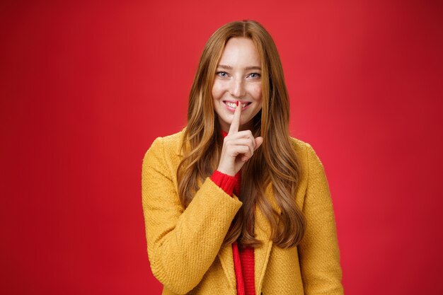 Chut, garde le secret. Fille au gingembre séduisante et douce, avec des taches de rousseur, des choses mystérieuses montrant un geste de chut avec l'index sur la bouche, souriant ayant une surprise sur fond rouge.