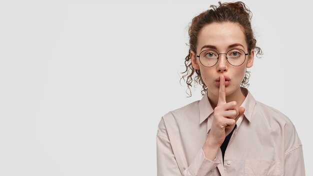 Chut, ne fais pas de bruit! Jolie femme sérieuse avec des taches de rousseur avec une expression confiante, montre un geste silencieux, demande à être silencieux, porte une chemise ample et des lunettes rondes, se dresse sur un mur blanc avec un espace vide