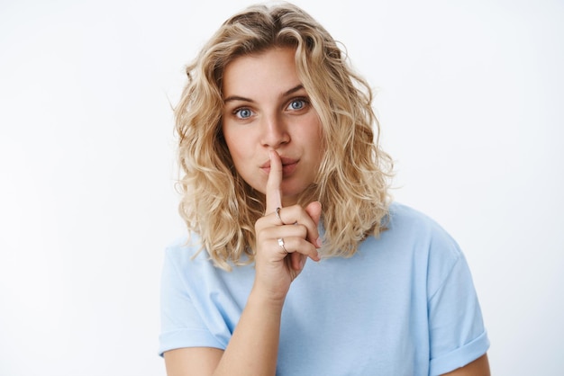 Chut pas de mots Portrait d'une séduisante jeune femme blonde européenne sensuelle et féminine aux yeux bleus et coupe de cheveux courte en t-shirt montrant le geste de chut avec l'index sur les lèvres