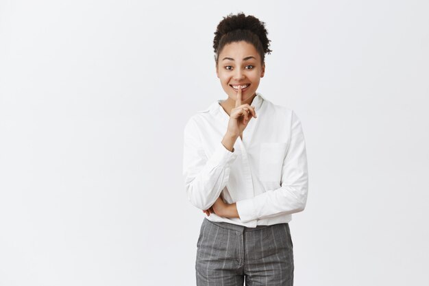 Chut, pas de mots s'il vous plaît. Portrait de charmante femme à la peau sombre et amicale aux cheveux bouclés en costume, gardant des secrets, montrant le geste chut avec l'index sur la bouche, souriant de bonheur