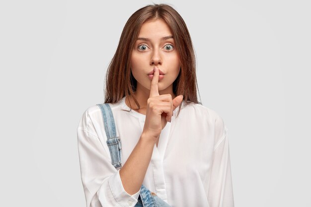 Chut, tais-toi! Une jolie femme sérieuse demande à garder les informations secrètes confidentielles, vêtue d'une chemise surdimensionnée blanche et d'une salopette en jean, pose contre un mur blanc. Concept de complot