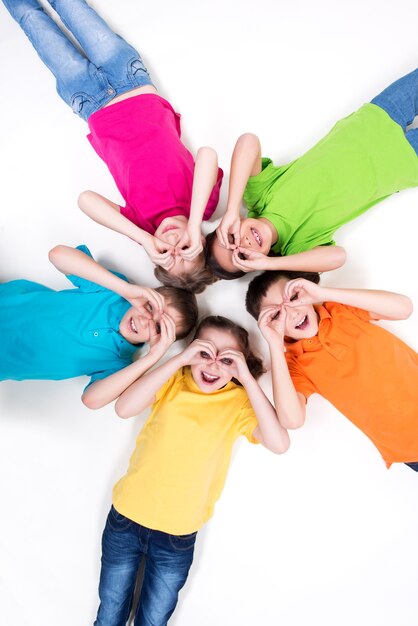 Cinq enfants heureux allongés sur le sol en cercle avec les mains près des yeux dans des t-shirts lumineux. Vue de dessus. Isolé sur blanc.