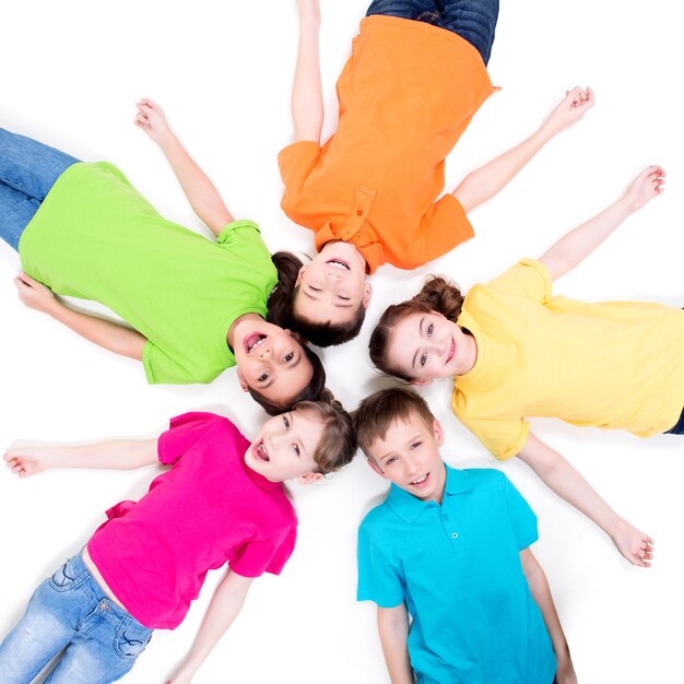 Cinq enfants souriants allongés sur le sol en cercle dans des t-shirts lumineux. Vue de dessus. Isolé sur blanc.