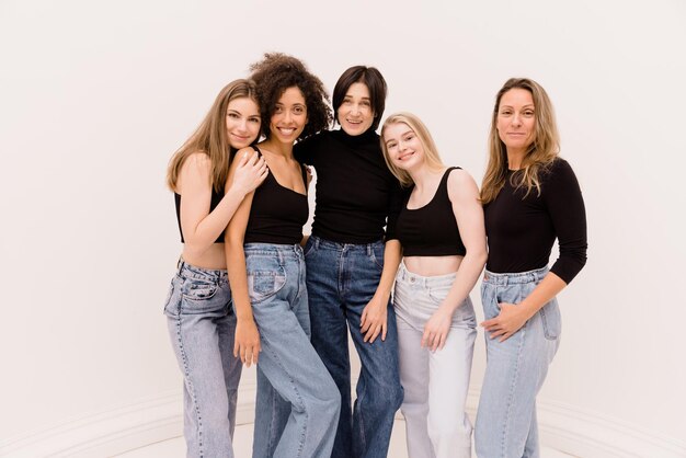 Cinq jeunes femmes adultes souriantes en jeans et pulls molletonnés se tiennent ensemble et regardent la caméra sur fond de studio blanc