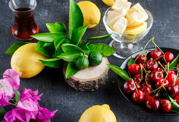 Citrons avec tranches, feuilles, verre de thé, fleurs, cerises, planche de bois sur une surface grise