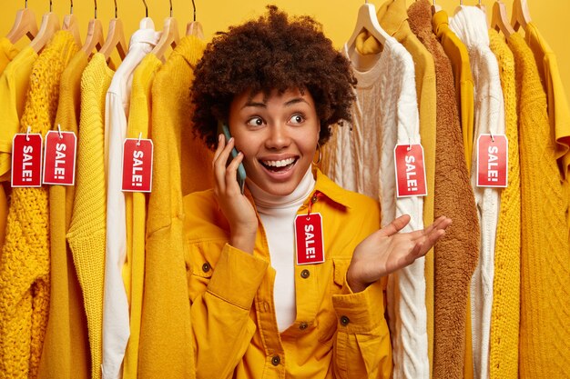 Une cliente heureuse a une conversation téléphonique avec un ami, demande des conseils sur ce qu'il faut mieux acheter, choisit une tenue dans un magasin de vêtements, aime faire du shopping, garde la paume de la main. Concept de vente au détail.