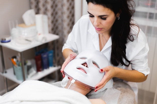 La cliente se trouve dans le salon sur la table de cosmétologie avec un masque blanc sur son visage