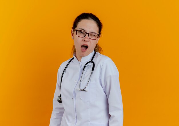 Clignotant jeune femme médecin portant une robe médicale et un stéthoscope avec des lunettes isolées