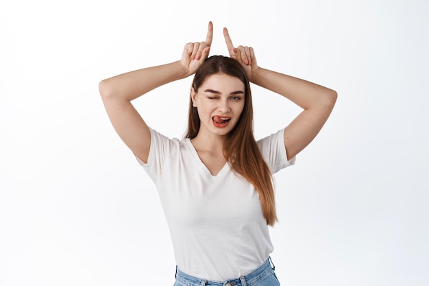 Clin d'œil impertinent et séduisant d'une jeune femme, montre le geste des cornes du diable de la langue et du taureau, les doigts sur la tête, s'affirmant et coquette, debout sur fond blanc