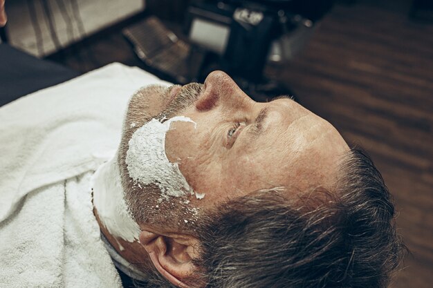 Close-up côté vue de dessus bel homme caucasien barbu senior se toilettage de barbe dans un salon de coiffure moderne.