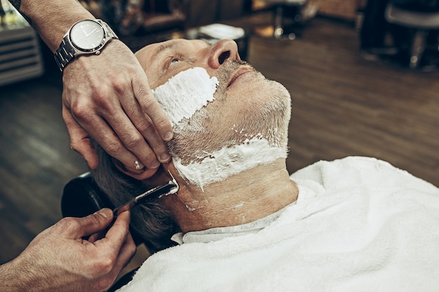 Close-up côté vue de dessus bel homme caucasien barbu senior se toilettage de barbe dans un salon de coiffure moderne.