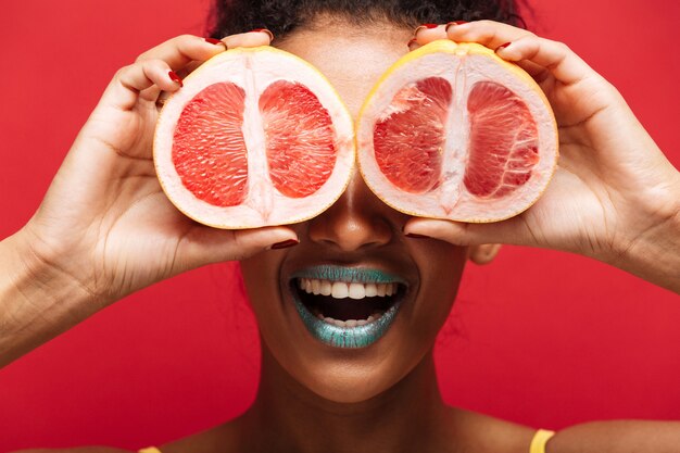 Close up food smiling afro american woman s'amuser couvrant les yeux avec deux moitiés de pamplemousse mûr frais, isolé sur mur rouge