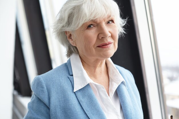 Close up image de l'élégant retraité femme d'âge moyen soigné avec des rides, des cheveux gris et un maquillage naturel debout à la fenêtre pendant la pause-café pendant la journée de travail au bureau, à la recherche