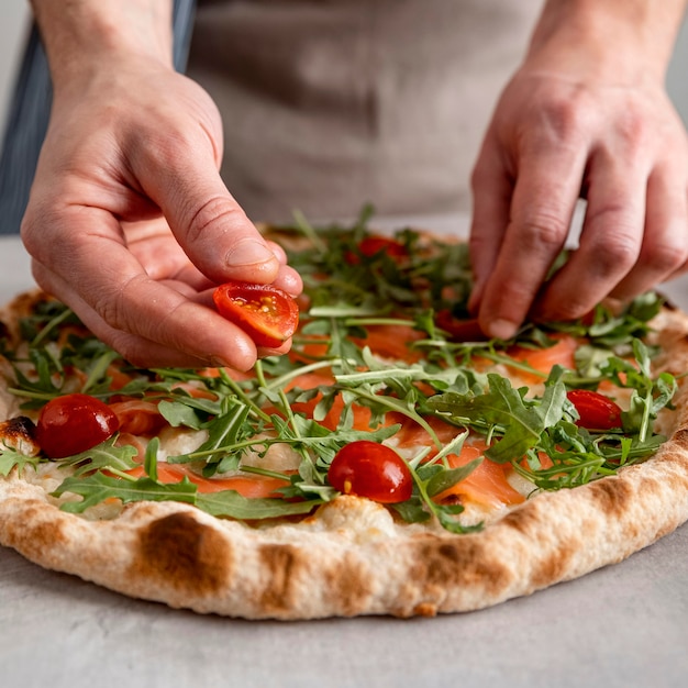 Photo gratuite close-up man putting tomates sur la pâte à pizza cuite au four avec des tranches de saumon fumé