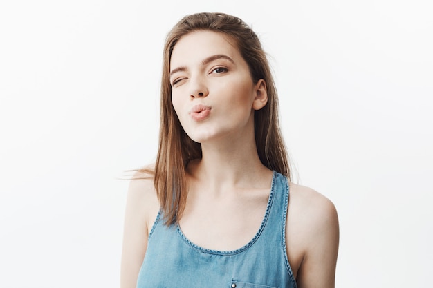 Close up of sexy attractive young European female collage student with dark hair in blue shirt winking,, making canard lips Posing for photo at friend's party.