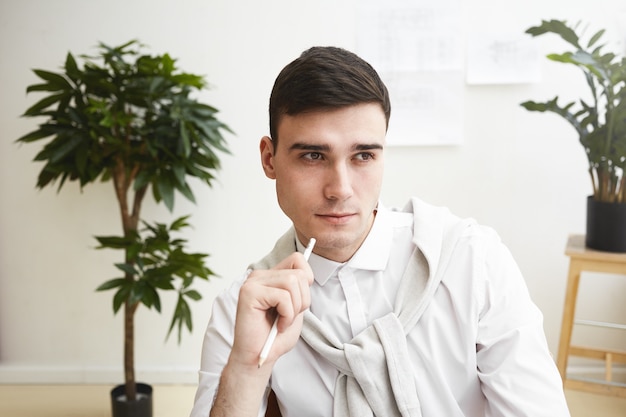 Close up portrait de beau jeune designer masculin européen rasé de près ayant un regard pensif tout en travaillant sur son lieu de travail, en pensant à de nouvelles idées et solutions. Les gens, le travail, le talent et la créativité