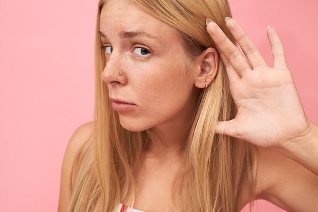 Close up Portrait de la belle adolescente blonde aux longs cheveux lâches, taches de rousseur et piercing nez plaçant la main à son oreille, ayant un regard curieux curieux